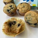 Blueberry lemon cornmeal muffins. four are visible in this photo, with one cut open so you can see the crumb. In the background is a pile of napkins in a fun blue and yellow print.