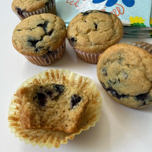 Blueberry lemon cornmeal muffins. four are visible in this photo, with one cut open so you can see the crumb. In the background is a pile of napkins in a fun blue and yellow print.